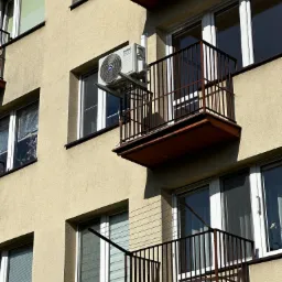 Zewnętrzna jednostka klimatyzatora Kaisai Eco zamontowana na ścianie bloku, widoczna z dołu, obok balkonu z brązową balustradą.