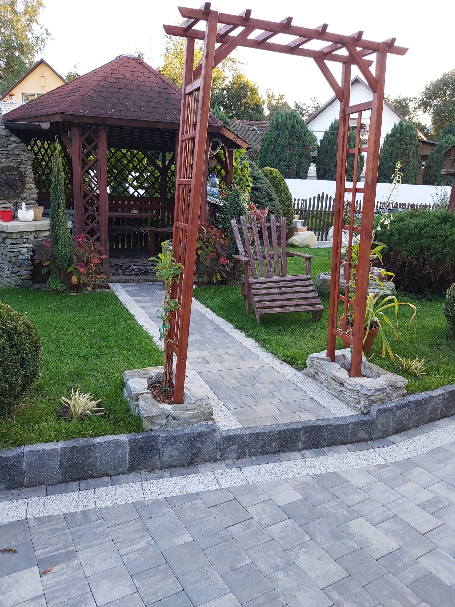 Ogród z altaną, kamienną ścieżką, drewnianą pergolą i meblami ogrodowymi, otoczony zielenią i roślinami ozdobnymi.