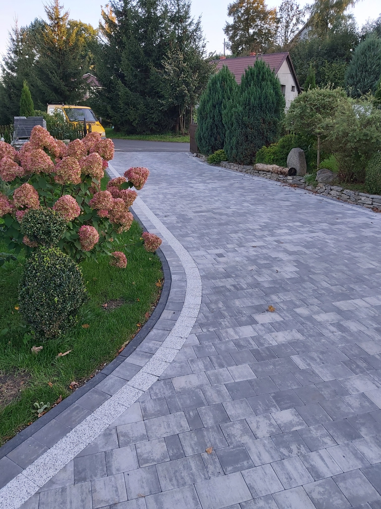 Szeroki podjazd z szarej kostki brukowej ułożonej we wzór, z obrzeżem z białego żwiru i rabatą z kwiatami hortensji, w tle widoczny żółty samochód dostawczy i dom.