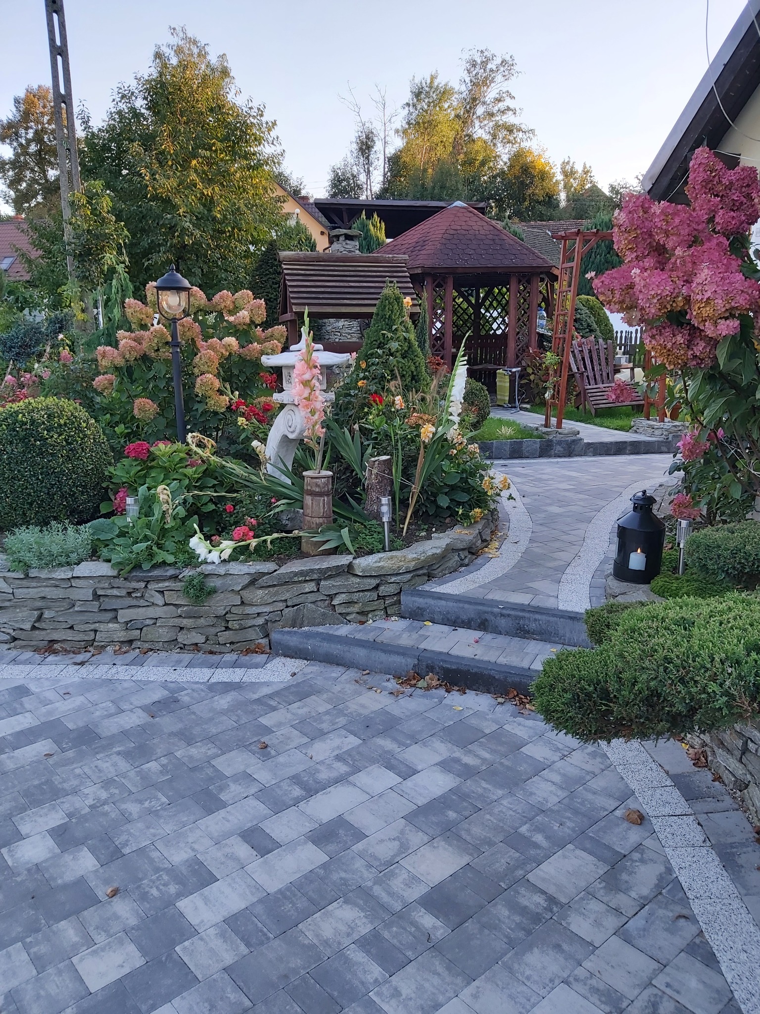Kaskadowa ścieżka z szarej kostki brukowej prowadząca przez zadbany ogród z gazebem, kwiatami, ozdobnymi krzewami i kamiennym murkiem oporowym.