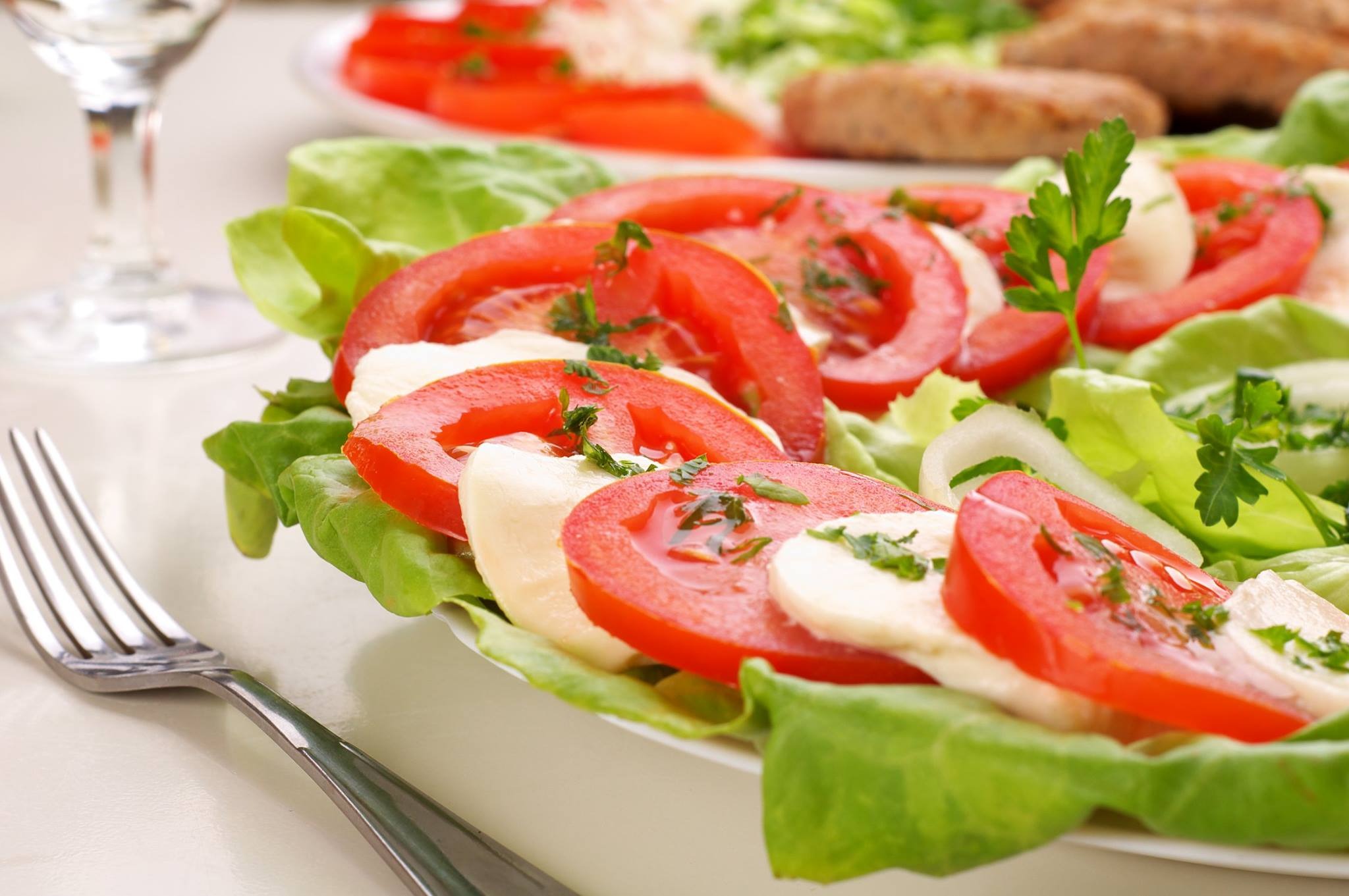 Caprese sałatka z plastrami pomidorów, mozzarelli i listkami bazylii, ułożona na zielonej sałacie, z widelcem obok na białym stole.