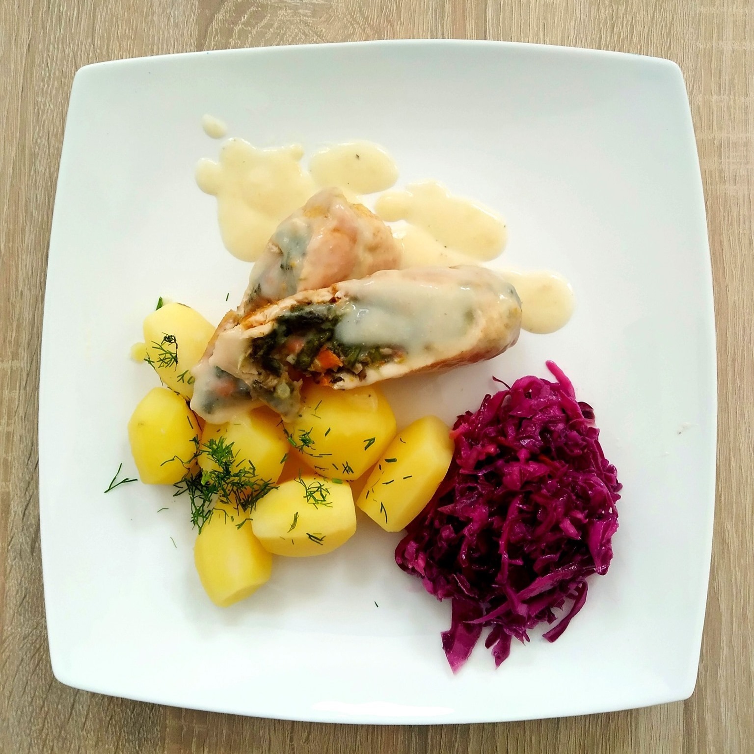 Rolada mięsna nadziewana warzywami, polana sosem, podana z gotowanymi ziemniakami posypanymi koperkiem i surówką z czerwonej kapusty na kwadratowym białym talerzu, całość ułożona na drewnianym blacie.