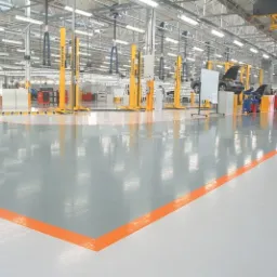 Błyszcząca, szara posadzka w hali produkcyjnej z wyznaczonymi pomarańczowymi liniami, widoczne podnośniki samochodowe i stanowiska pracy.