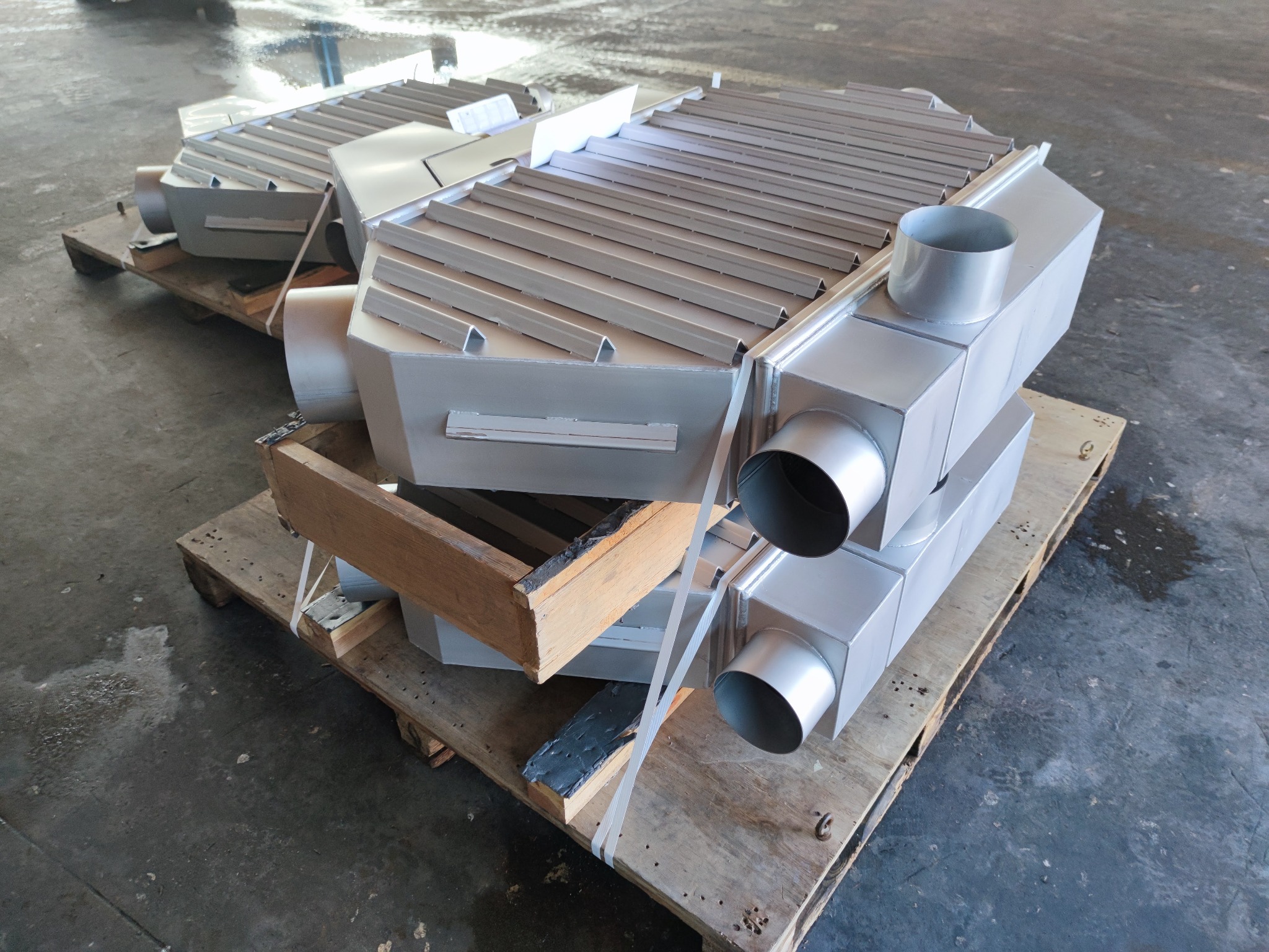 Dwa metalowe wymienniki ciepła z rurami, świeżo pomalowane na srebrno, ułożone na drewnianych paletach w hali produkcyjnej, widoczne ślady spawania.