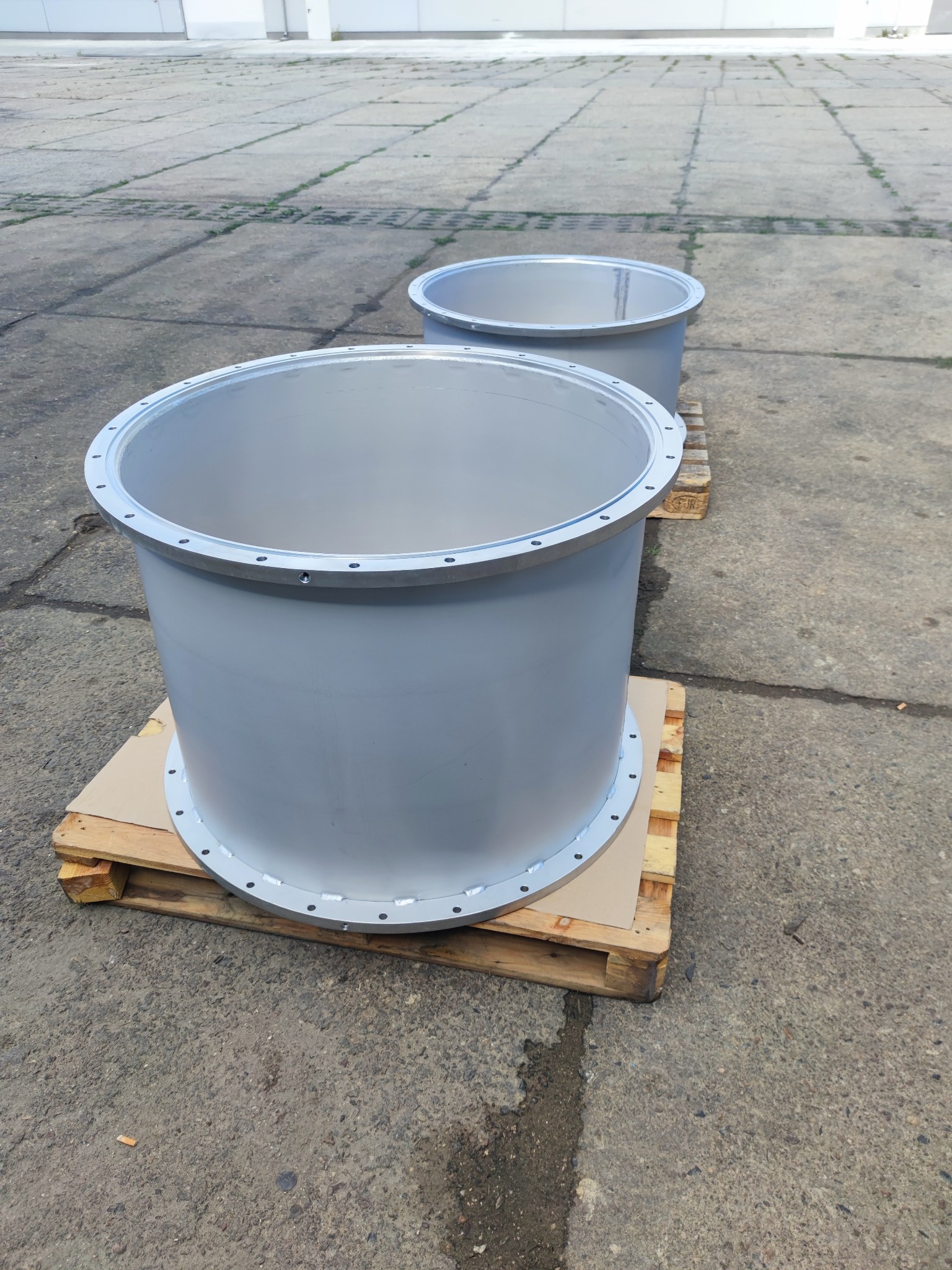 Dwa szare, metalowe cylindry z kołnierzami na paletach, widoczne ślady spawania i mocowania śrubami, na tle betonowego placu.