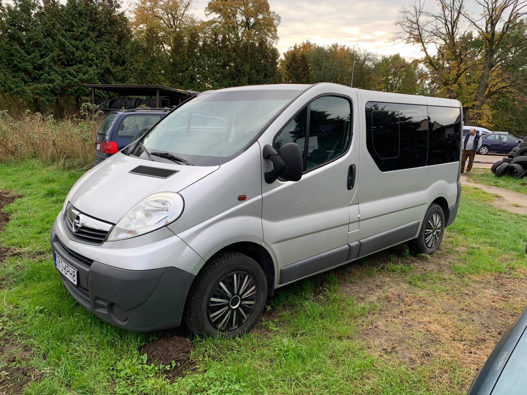 Srebrny Opel Vivaro 9-osobowy z 2013 roku, zaparkowany na trawiastym terenie, z widocznymi oponami w tle i osobą stojącą obok.