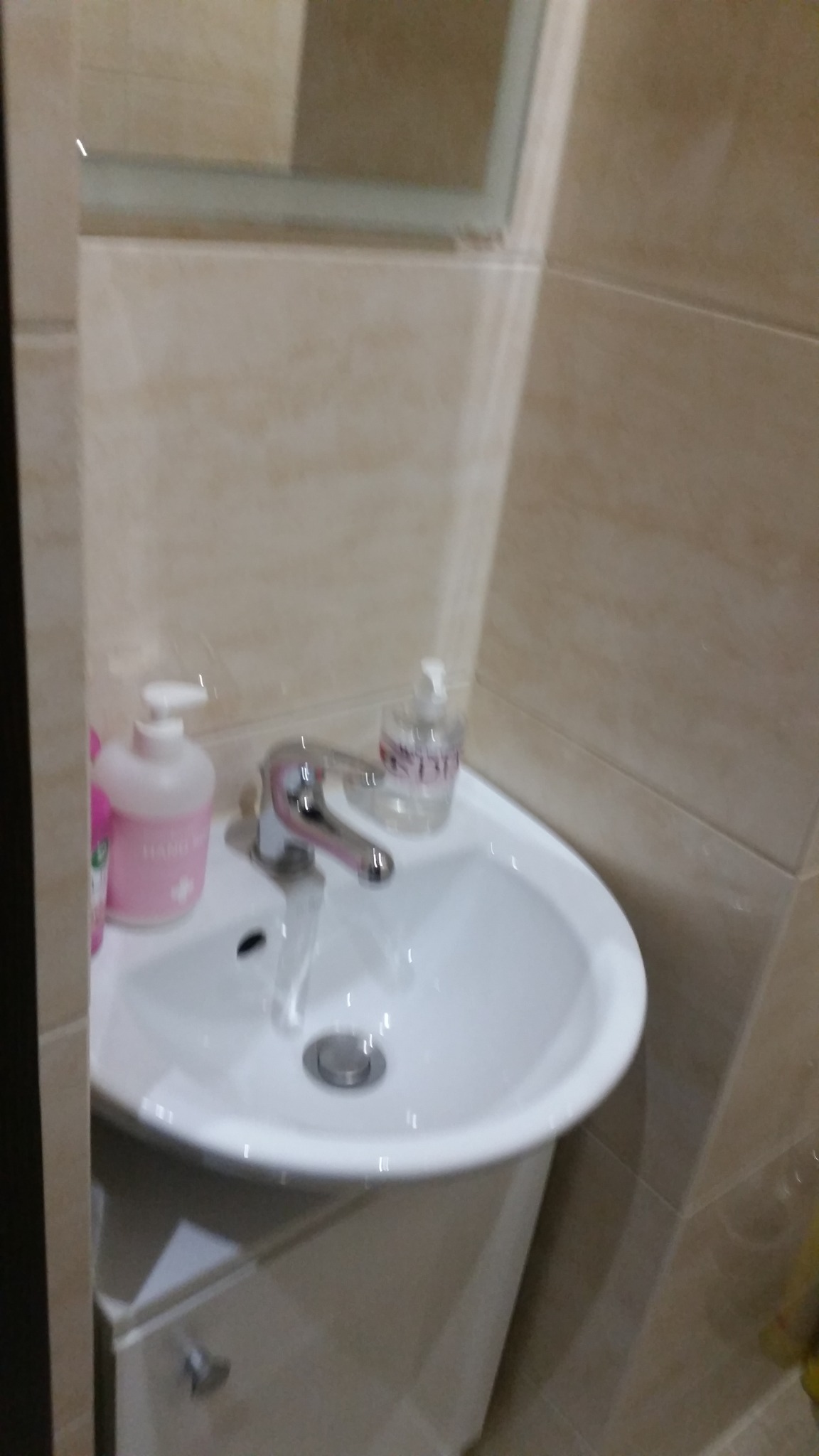 Biała umywalka z baterią, na blacie dwie butelki z mydłem w dozownikach, beżowe płytki na ścianie i szafka pod umywalką.