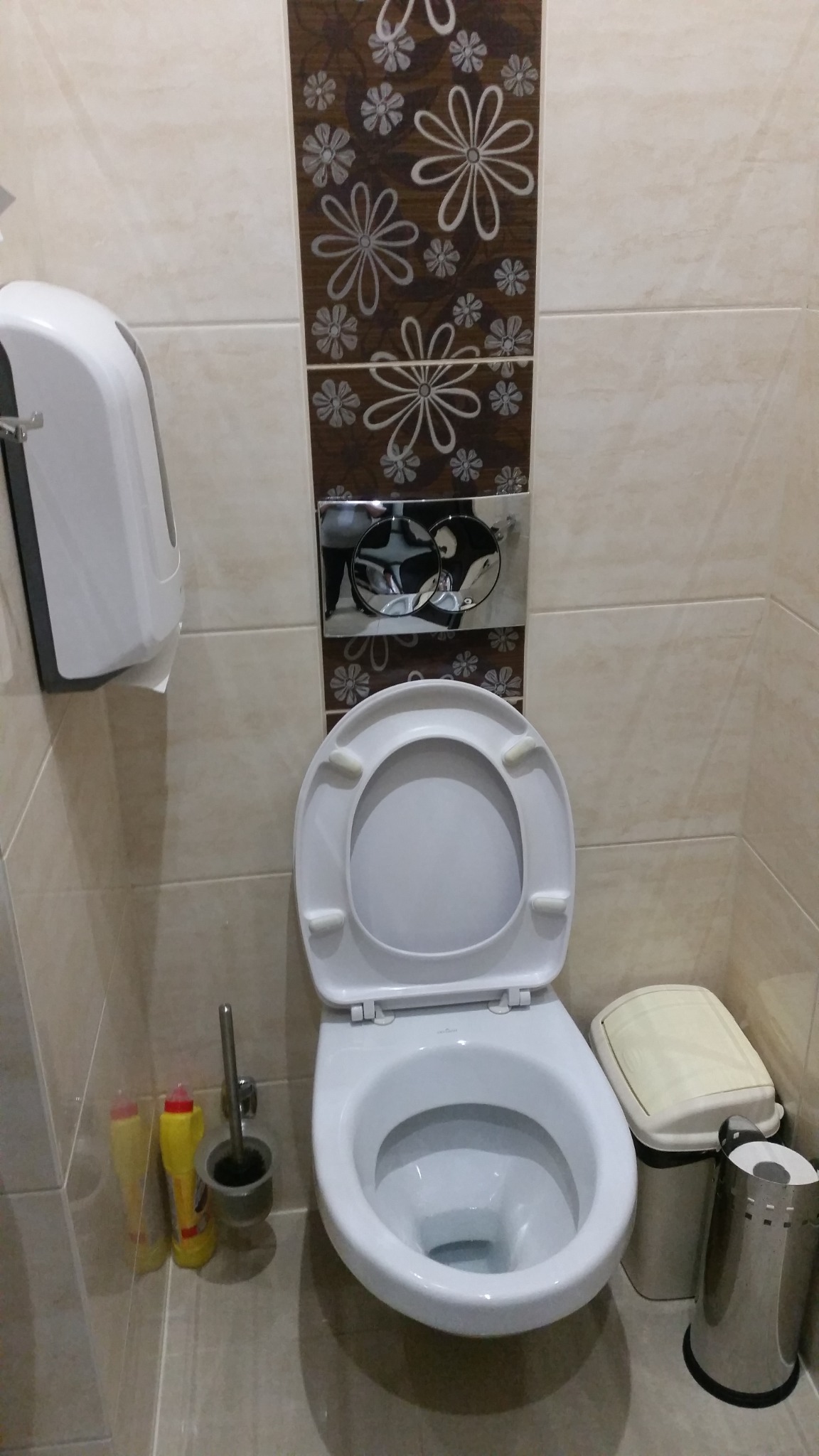 Toaleta z otwartą klapą w łazience z beżowymi płytkami i dekoracyjnym pasem z motywem kwiatowym, obok kosz na śmieci i szczotka do WC.