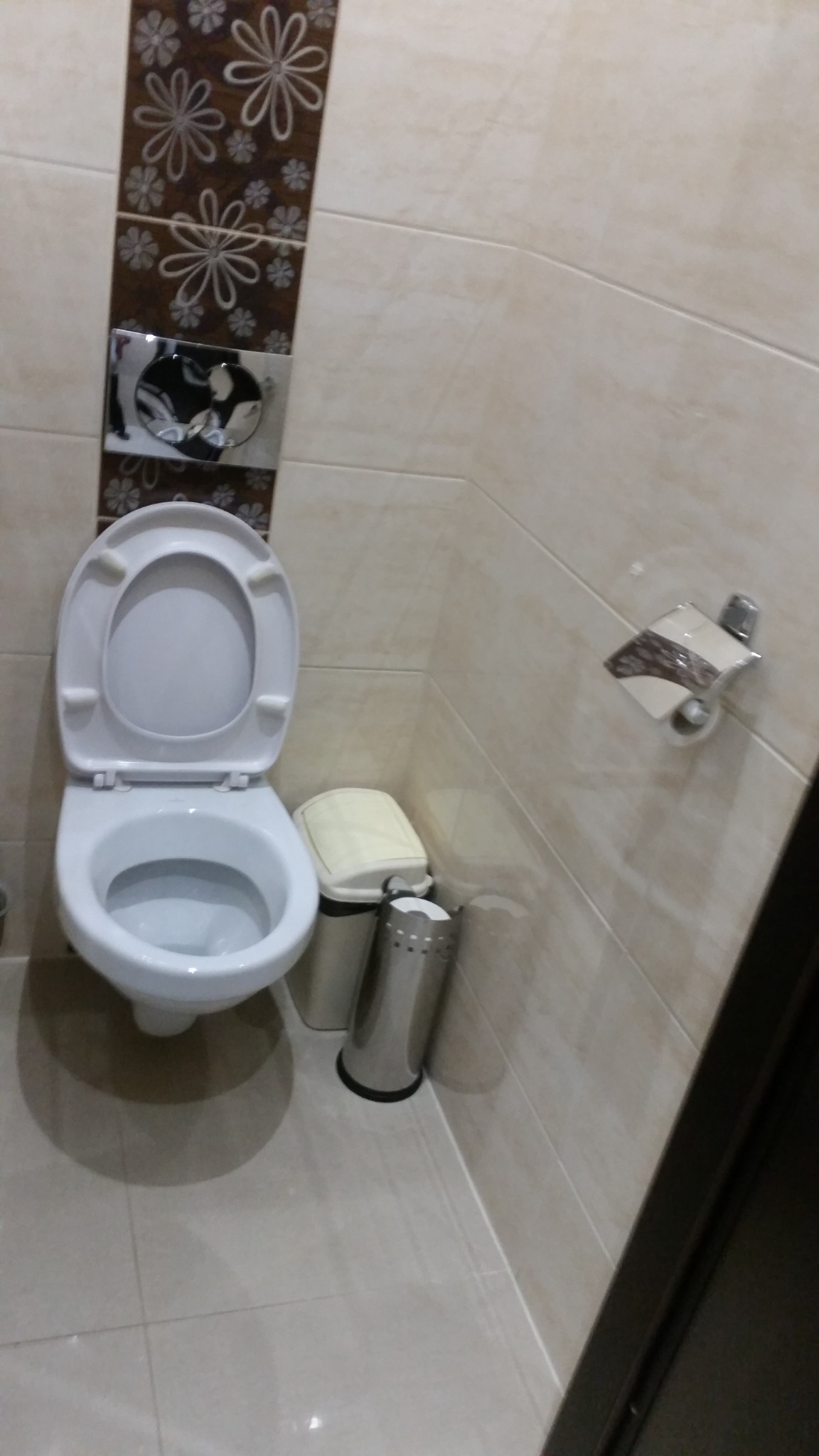 Toaleta z otwartą klapą, obok kosze na śmieci i uchwyt na papier toaletowy na tle beżowych płytek z dekoracyjnym pasem z motywem kwiatowym.