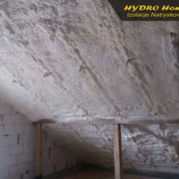 Poddasze z widoczną białą, natryskową izolacją piankową na skośnym dachu i ścianach z bloczków, z drewnianymi belkami podtrzymującymi.