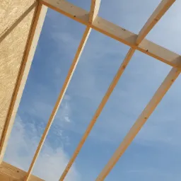 Surowa konstrukcja drewniana domu w budowie widziana od wewnątrz, z widokiem na błękitne niebo z chmurami przez otwory w dachu.