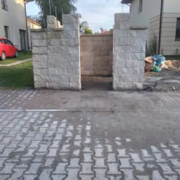 Wznoszenie bramy wjazdowej z bloków betonowych o nieregularnej fakturze, widoczne fragmenty konstrukcji i otoczenie placu budowy.