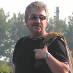 Portret mężczyzny w okularach przeciwsłonecznych, z kurtką na ramieniu, na tle zieleni i komina przemysłowego.