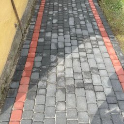 Para-System - Ułożona kostka brukowa w odcieniach szarości z czerwonym obramowaniem, widoczne ślady wilgoci po niedawnym czyszczeniu, zarys budynku w tle.