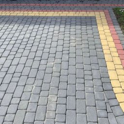 Para-System - Ułożona szara kostka brukowa z bordiurą w kolorze żółtym i czerwonym przy ścianie budynku z czerwonymi drzwiami.