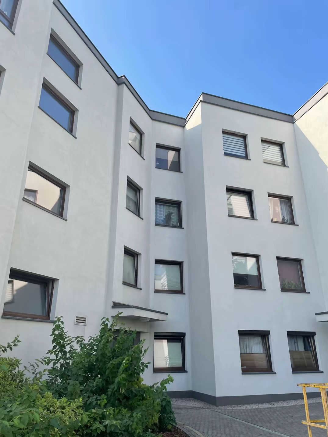 Wielopiętrowy budynek z jasną elewacją, widok z dołu na fragment fasady z licznymi oknami i zielenią u podstawy.