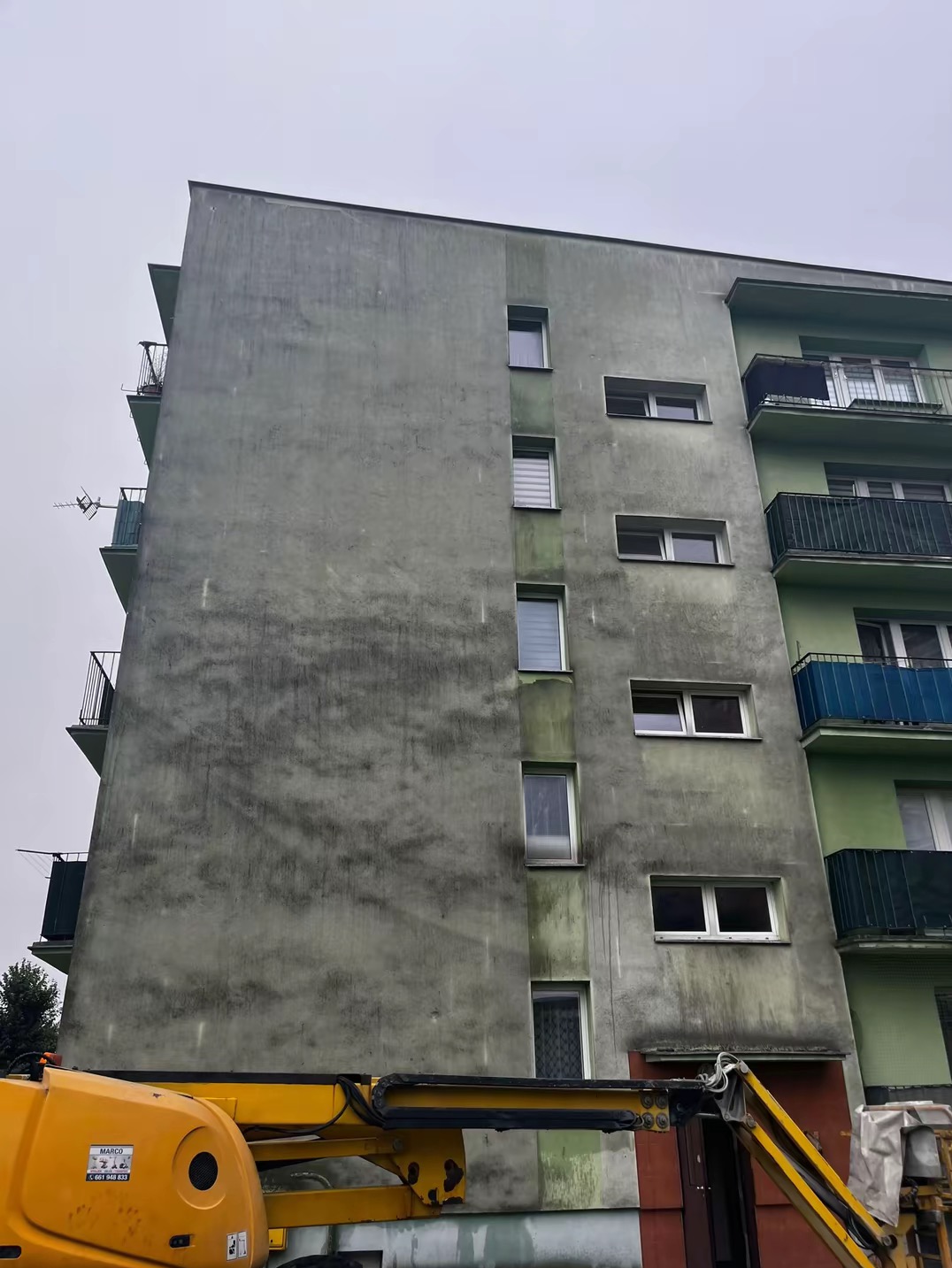 Wielopiętrowy budynek mieszkalny z widoczną różnicą w czystości elewacji, gdzie część jest umyta, a część jeszcze zabrudzona. Na pierwszym planie fragment żółtego podnośnika koszowego.