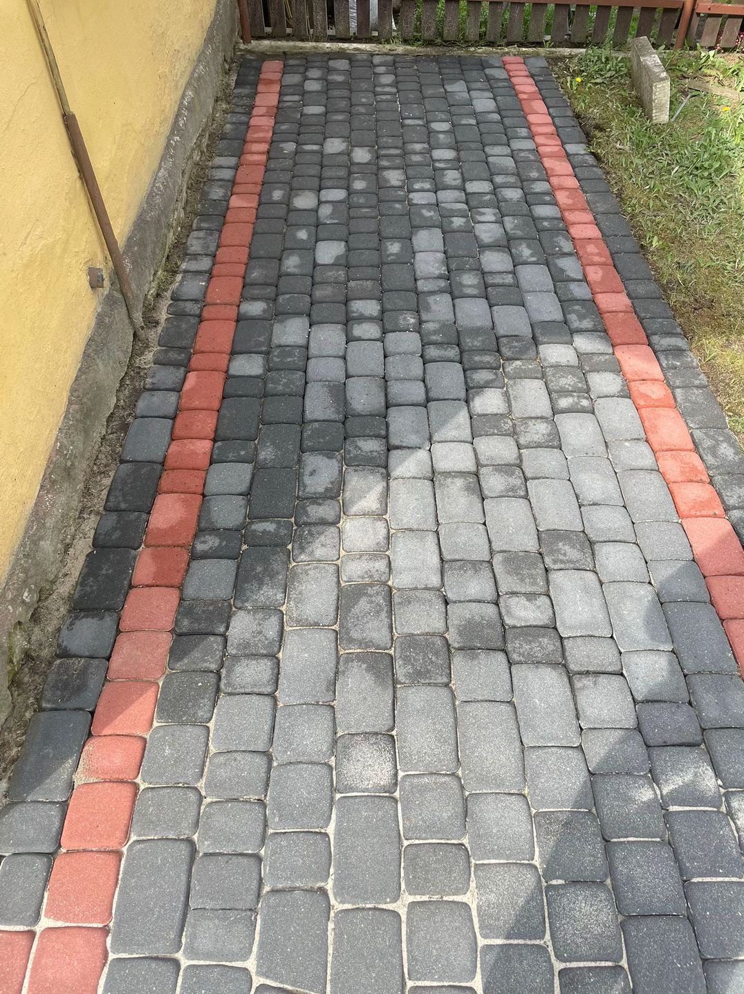 Ułożona kostka brukowa w odcieniach szarości z czerwonym obramowaniem, widoczne ślady wilgoci po niedawnym czyszczeniu, zarys budynku w tle.