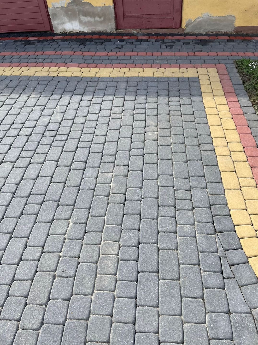 Ułożona szara kostka brukowa z bordiurą w kolorze żółtym i czerwonym przy ścianie budynku z czerwonymi drzwiami.