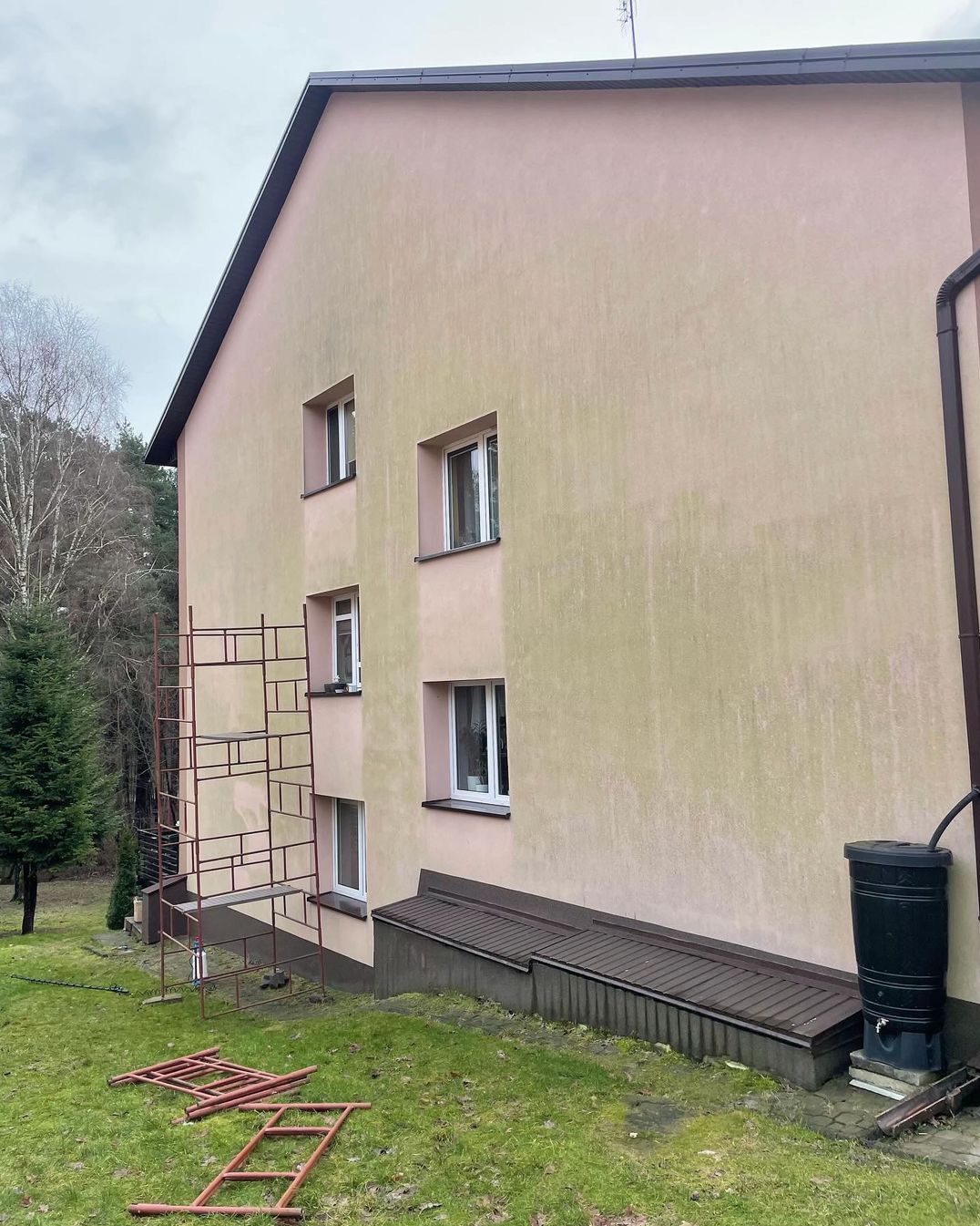 Budynek z elewacją pokrytą zielonym nalotem, częściowo zasłonięty przez metalowe rusztowanie, z czarnym zbiornikiem na deszczówkę przy ścianie.