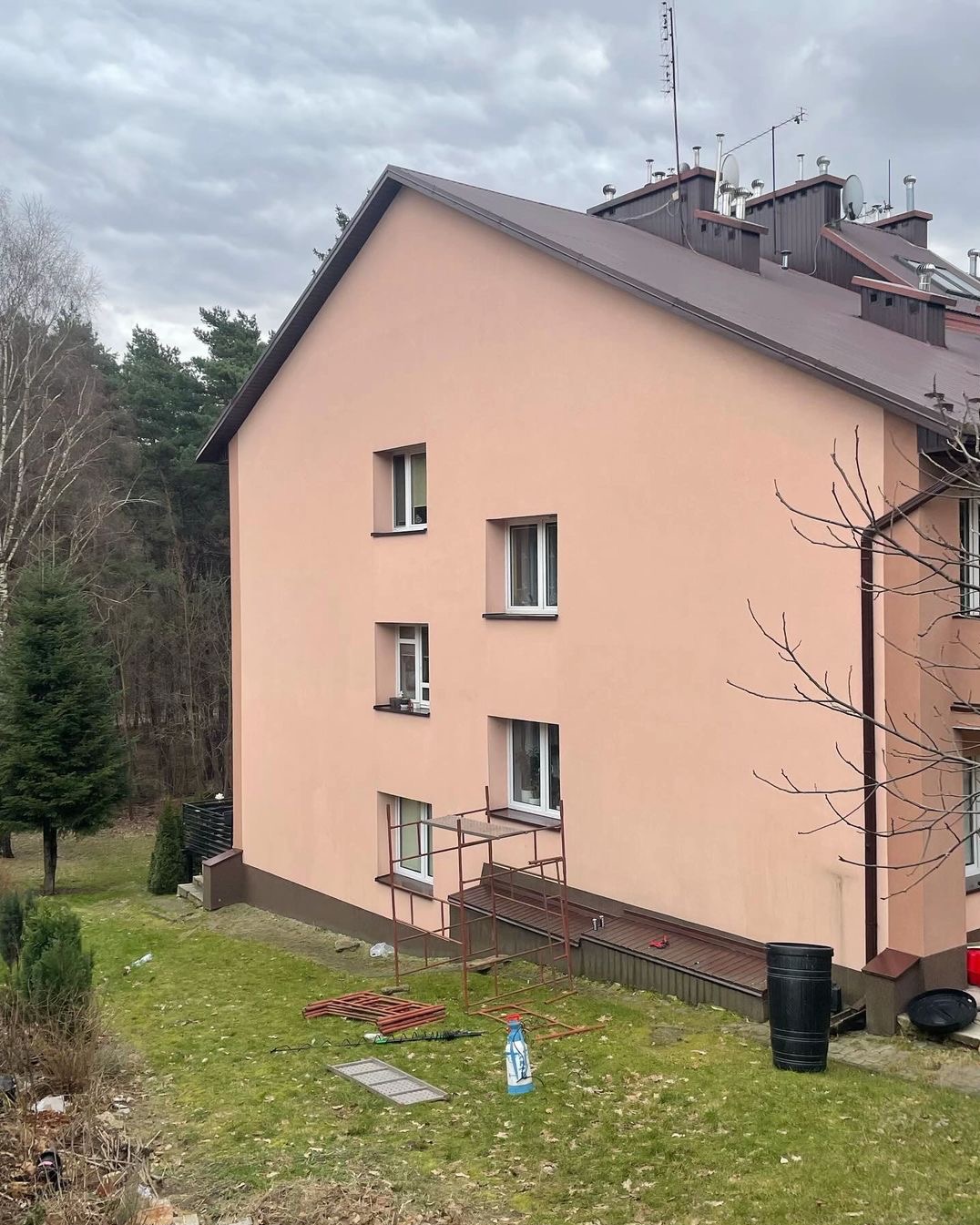 Dwupiętrowy budynek z różową elewacją, widoczne rusztowanie przy oknie na parterze, butelka środka czyszczącego i fragmenty rusztowania na trawniku, pochmurne niebo w tle.