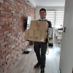 Młody mężczyzna w roboczym stroju i czapce niesie dwie kartonowe paczki w korytarzu z ceglaną ścianą, widoczny odkurzacz i stół z krzesłami w tle.