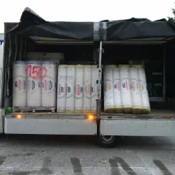 Otwarta paka białego busa wypełniona rolkami izolacji Ursa Glasswool Amber 33 i Gold 35 na paletach, z czerwoną liczbą '150' namalowaną na jednej z nich, na tle zimowego krajobrazu.