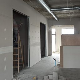 Fach-remont - Prace wykończeniowe w biurze: pracownik na drabinie montuje płyty gipsowo-kartonowe, widoczne profile metalowe, częściowo wyszpachlowane ściany, instalacje pod sufitem.