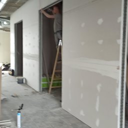 Fach-remont - Pracownik na drabinie montuje płytę gipsowo-kartonową w nowo powstającym pomieszczeniu biurowym z widocznymi instalacjami wentylacyjnymi pod sufitem.