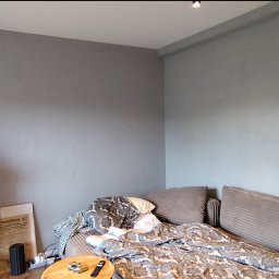 Fach-remont - Szara ściana z efektem dekoracyjnym w pokoju, widok na fragment sofy z poduszkami, roślina doniczkowa i gazeta. Minimalistyczne wnętrze po malowaniu.