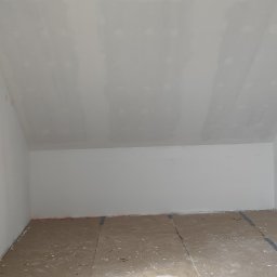 Fach-remont - Poddasze po gipsowaniu, widoczne łączenia płyt i zabezpieczona podłoga. W tle białe ściany i sufit ze skosem. Wystający kabel z sufitu.