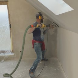 Fach-remont - Pracownik w nausznikach szlifuje ścianę na poddaszu szlifierką z odkurzaczem, przygotowując ją do dalszych prac wykończeniowych w nowym mieszkaniu.