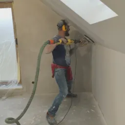 Pracownik w nausznikach szlifuje ścianę na poddaszu szlifierką z odkurzaczem, przygotowując ją do dalszych prac wykończeniowych w nowym mieszkaniu.