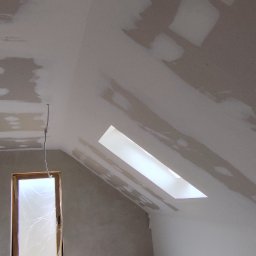 Fach-remont - Poddasze w trakcie prac wykończeniowych: widoczne płyty karton-gips z nałożoną warstwą szpachli. Okno dachowe i pionowe w tle. Kabel zwisa ze stropu.
