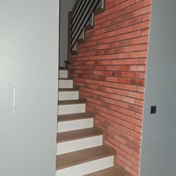 Fach-remont - Schody z drewnianymi stopniami i białą podstopnicą, obok ściana wyłożona czerwoną cegłą. Szara ściana z lewej strony kadru.