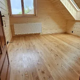 Jasne drewniane wnętrze poddasza z oknami: standardowym i dachowym. Widoczne deski na ścianach, podłodze i suficie, grzejnik pod oknem. Naturalne wykończenie.