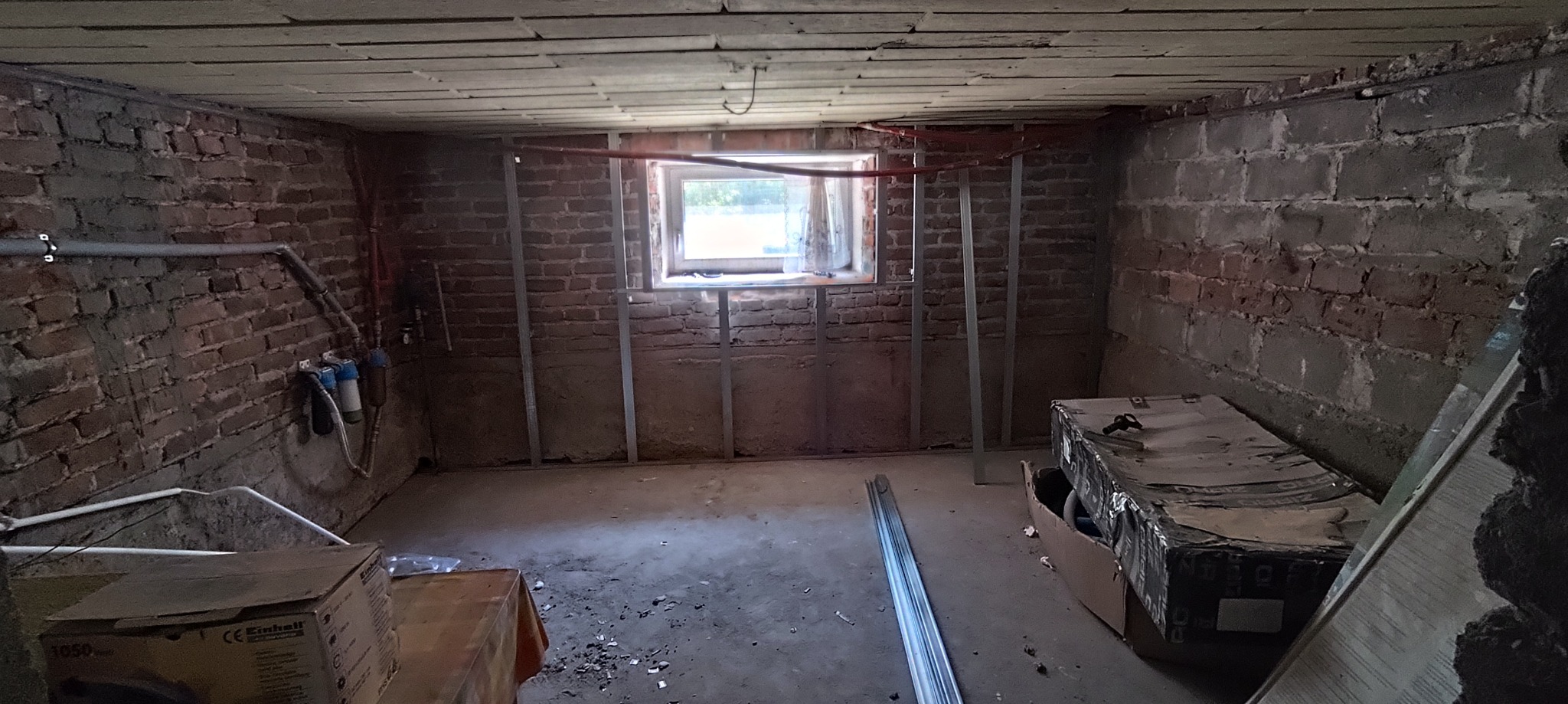 Piwnica w trakcie remontu: surowe cegły, metalowe profile, instalacje wodne, okno. Przygotowanie do prac wykończeniowych, widoczne materiały budowlane.