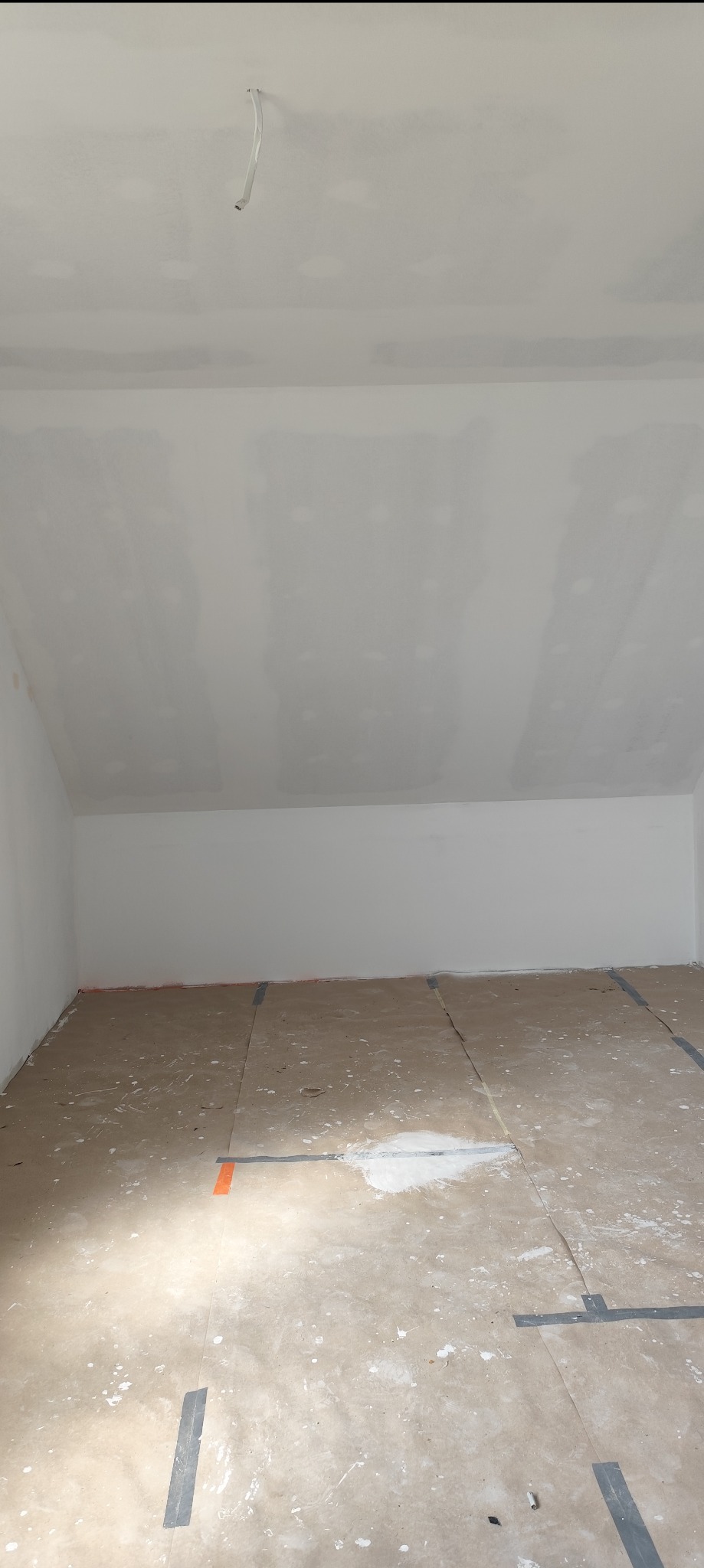 Poddasze po gipsowaniu, widoczne łączenia płyt i zabezpieczona podłoga. W tle białe ściany i sufit ze skosem. Wystający kabel z sufitu.