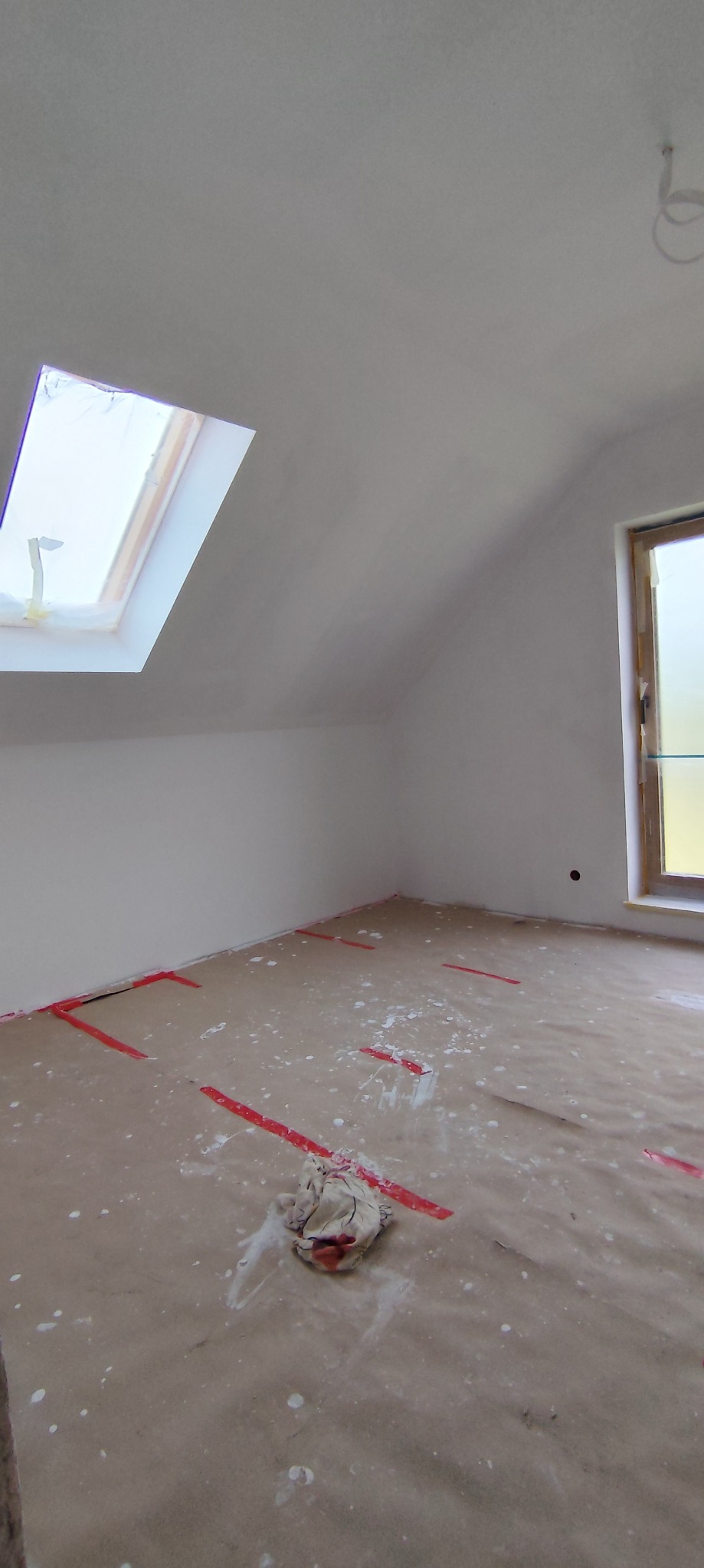 Poddasze po gładziowaniu ścian, z oknem dachowym i zabezpieczoną podłogą. Widoczne ślady gładzi na podłodze i taśma ochronna przy listwach.