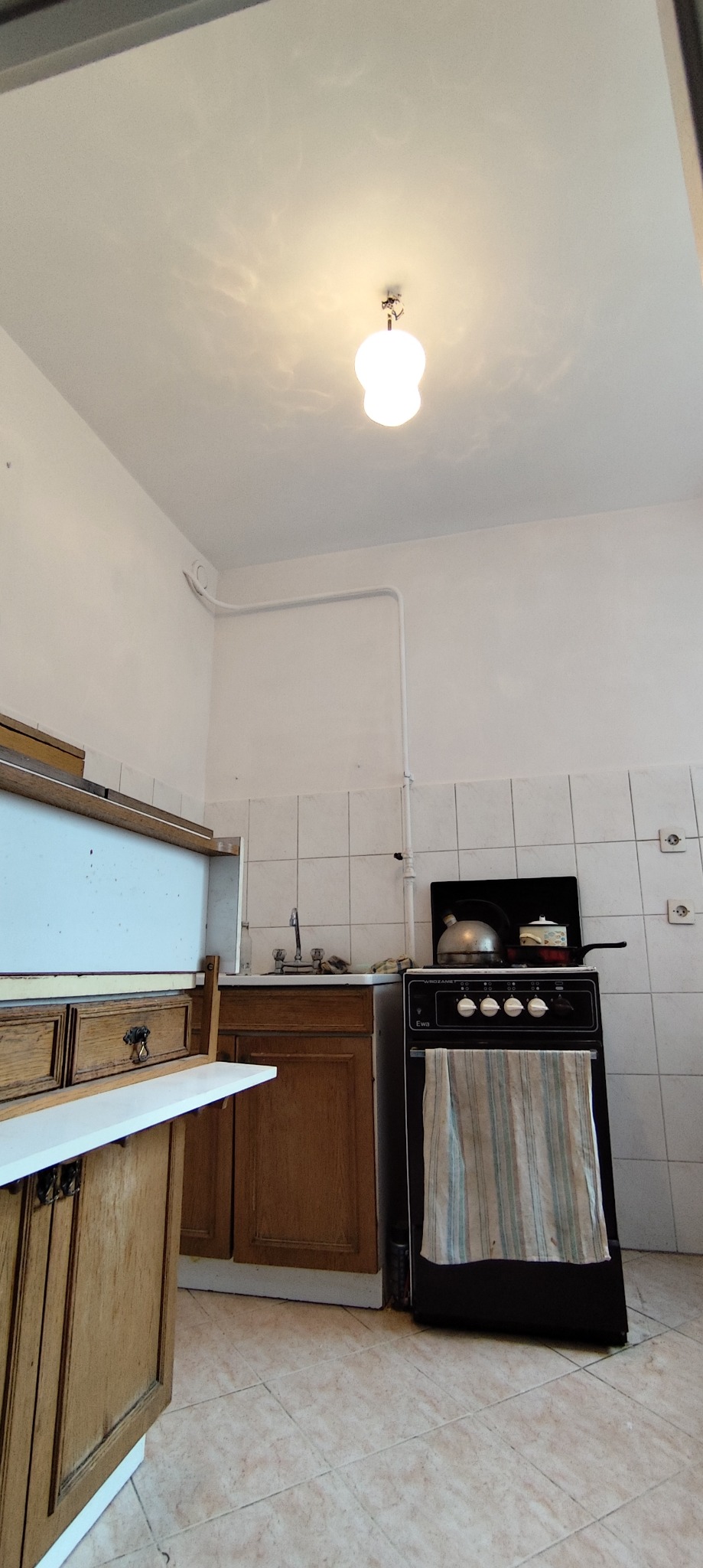 Kuchnia z lat 80.: drewniane szafki, białe płytki, czarna kuchenka z czajnikiem. Widoczne gniazdka i rury na ścianie. Ujęcie wnętrza w trakcie użytkowania.