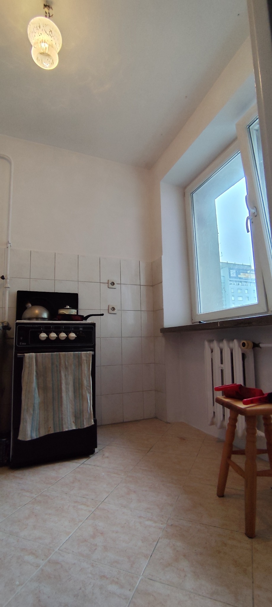 Kuchnia przed remontem: stara kuchenka z garnkami, okno z widokiem na blok, ceramiczne płytki na ścianach i podłodze, drewniany taboret. Wnętrze wymaga odświeżenia.