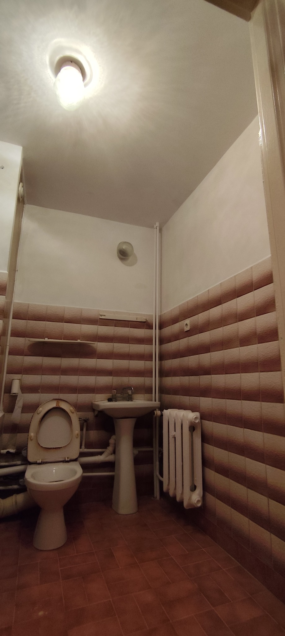 Ujęcie toalety z umywalką na postumencie i grzejnikiem, wykończonej brązowymi, poziomymi płytkami. Widoczne elementy instalacji i oświetlenie.