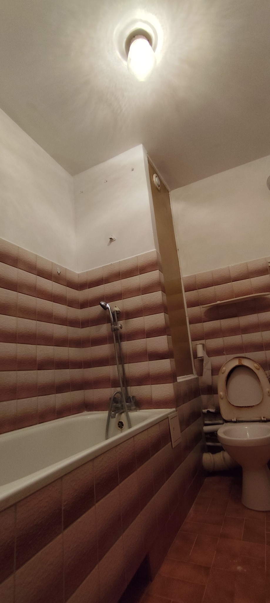 Łazienka z brązowo-białymi płytkami, wanną i toaletą. Widoczne niedoskonałości i ślady użytkowania. Ujęcie z dołu, widoczna lampa na suficie.