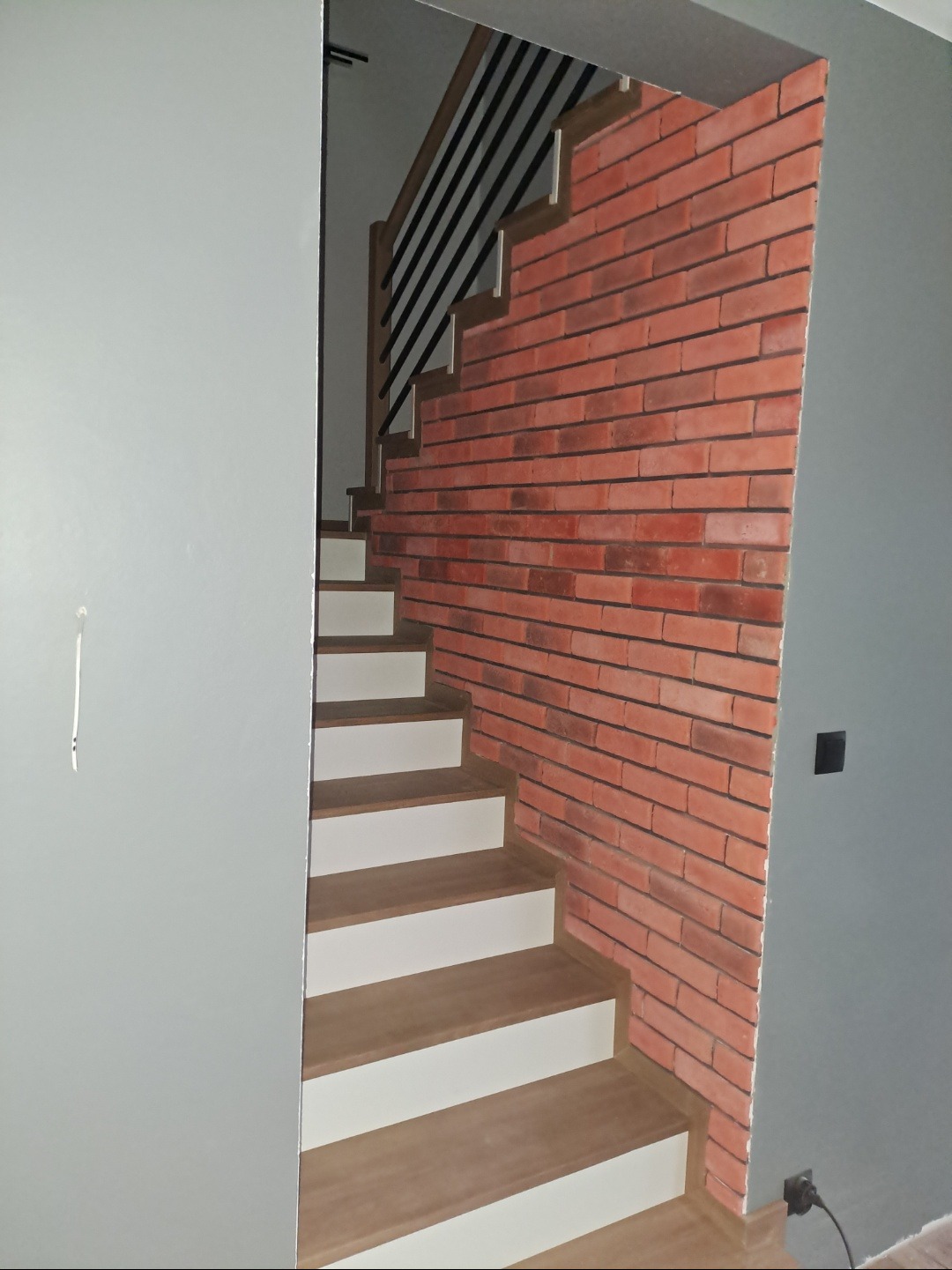 Schody z drewnianymi stopniami i białą podstopnicą, obok ściana wyłożona czerwoną cegłą. Szara ściana z lewej strony kadru.