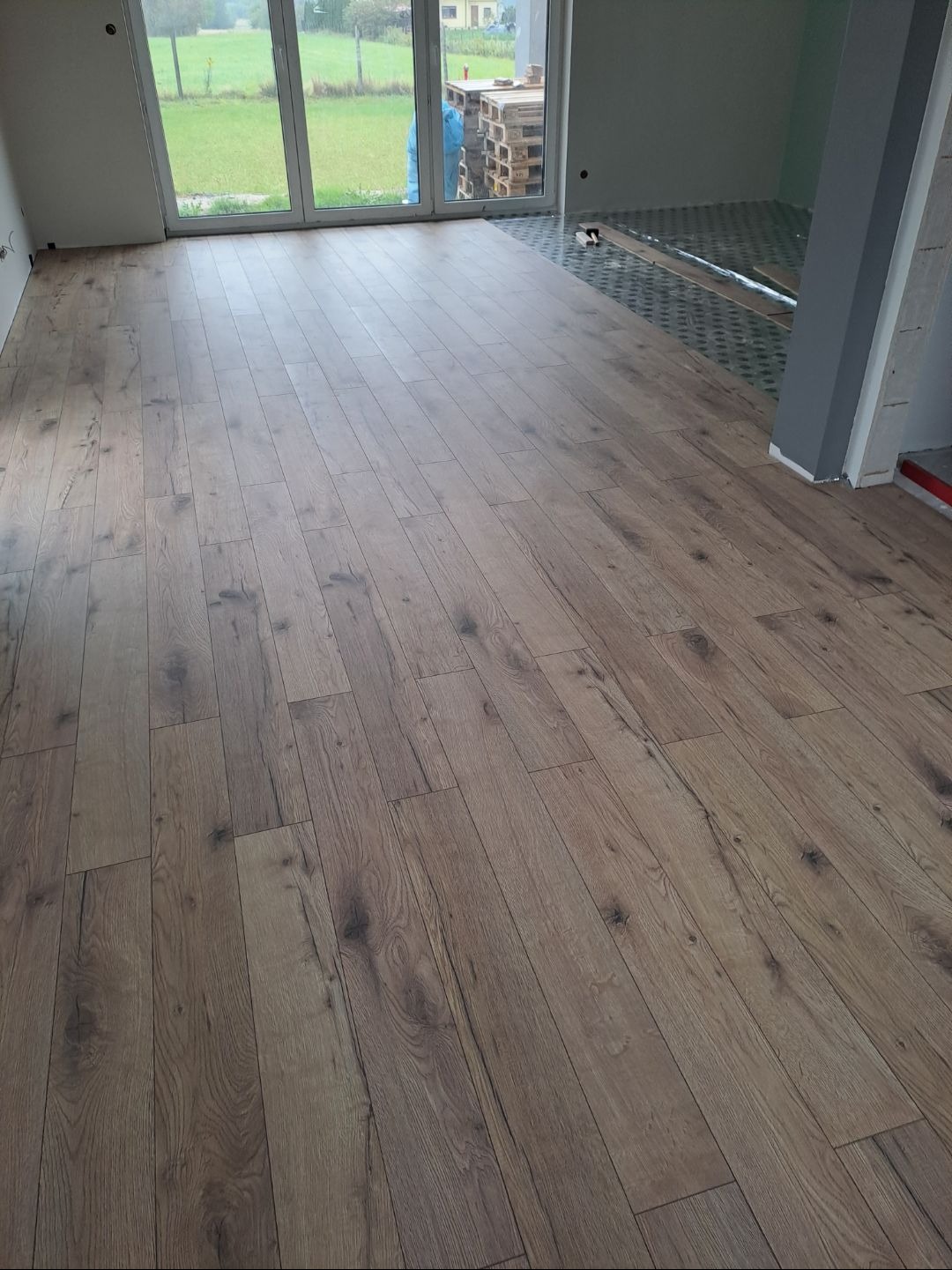 Ukończona podłoga z paneli w odcieniach brązu, widok na duże okno z zielonym krajobrazem. Część podłogi jeszcze nieukończona z widoczną izolacją.