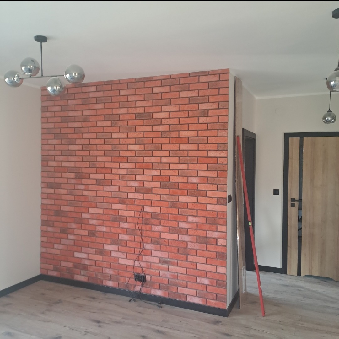 Ściana z cegły dekoracyjnej w nowoczesnym wnętrzu, z widocznymi kablami i poziomnicą, oświetlona przez designerską lampę sufitową.
