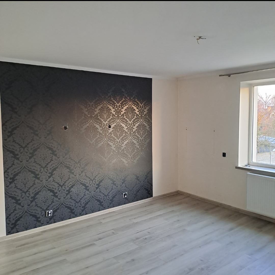 Pokój po remoncie: ściana z wzorzystą, czarną tapetą, jasne ściany, szara podłoga. Widoczne gniazdka i okno z grzejnikiem. Minimalistyczny, nowoczesny design.