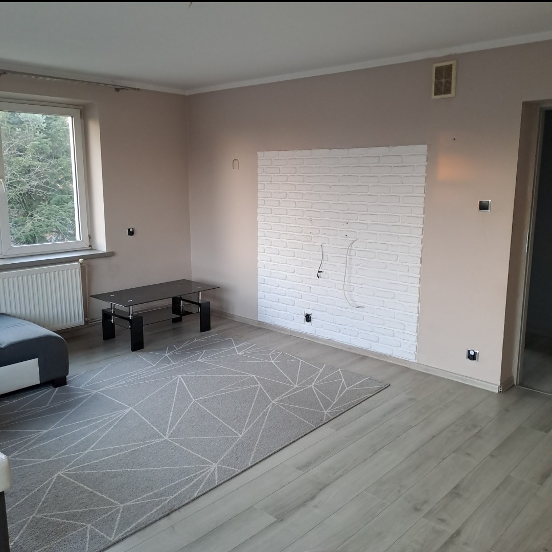 Jasny salon po remoncie: ściana z białej cegły, szary dywan w geometryczne wzory, drewniana podłoga i beżowe ściany. Widoczne gniazdka i wyprowadzone kable.