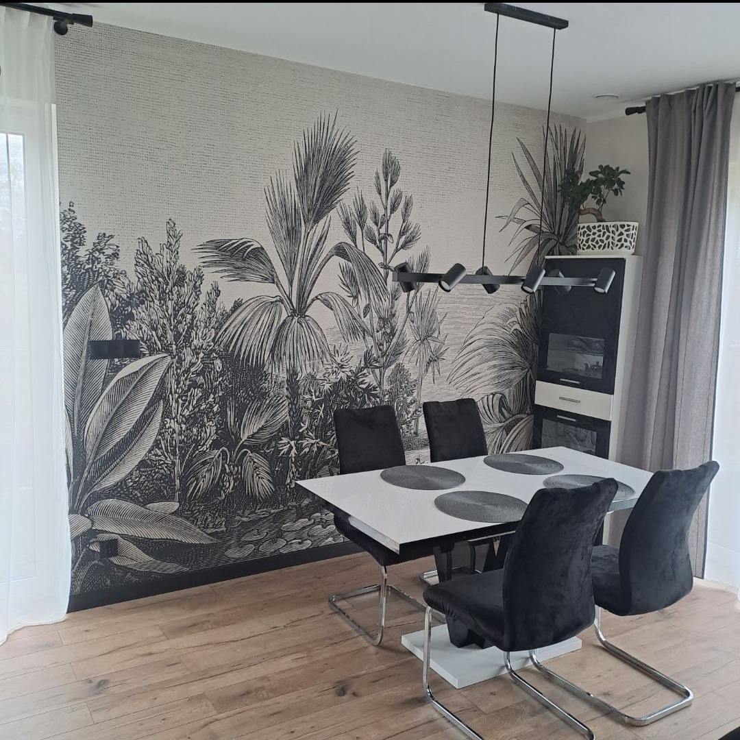 Jadalnia z fototapetą w stylu dżungli, czarne krzesła i biały stół. Nowoczesne oświetlenie sufitowe. Parkiet na podłodze.