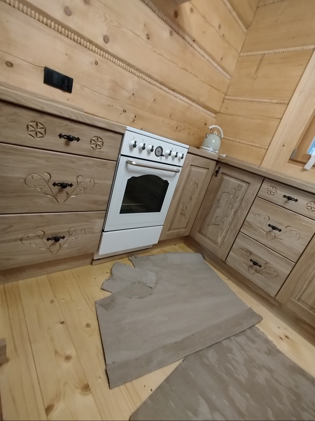 Kuchnia w stylu rustykalnym z drewnianymi szafkami zdobionymi rzeźbieniami, białą kuchenką i fragmentami zabezpieczeń podłogi podczas prac wykończeniowych.